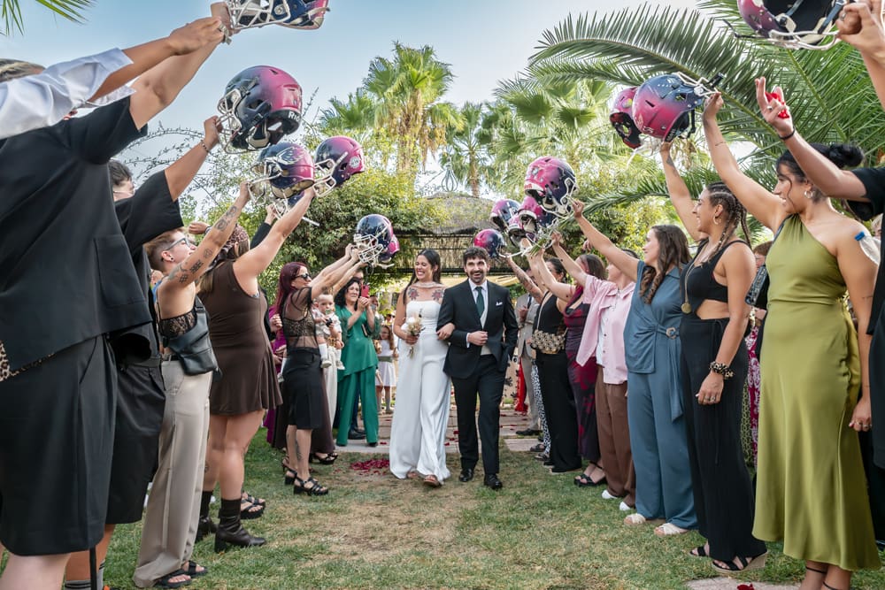Boda civil original en Hotel Boabdil Granada. Los invitados levantan cascos personalizados en un pasillo de honor para Jose y Cristina, que sonríen entre ellos. Un momento divertido capturado por fotógrafo bodas Granada.