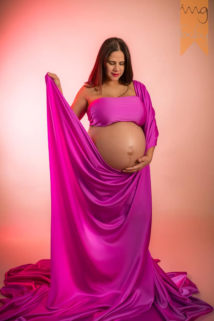 Mujer embarazada posando en una sesión de fotos de maternidad, envuelta en una elegante tela de satén color magenta sobre un fondo rosado suave.