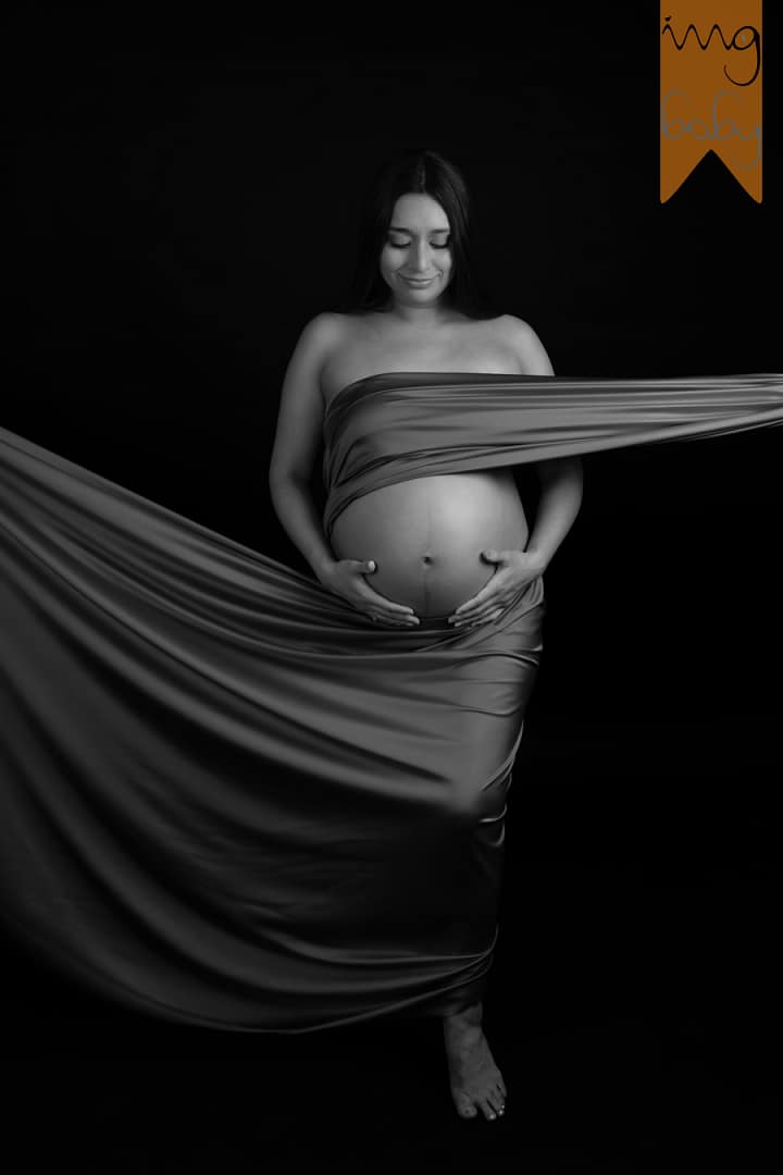 Fotografía en blanco y negro de estudio de mujer embarazada envuelta en una tela fluida sobre fondo negro.